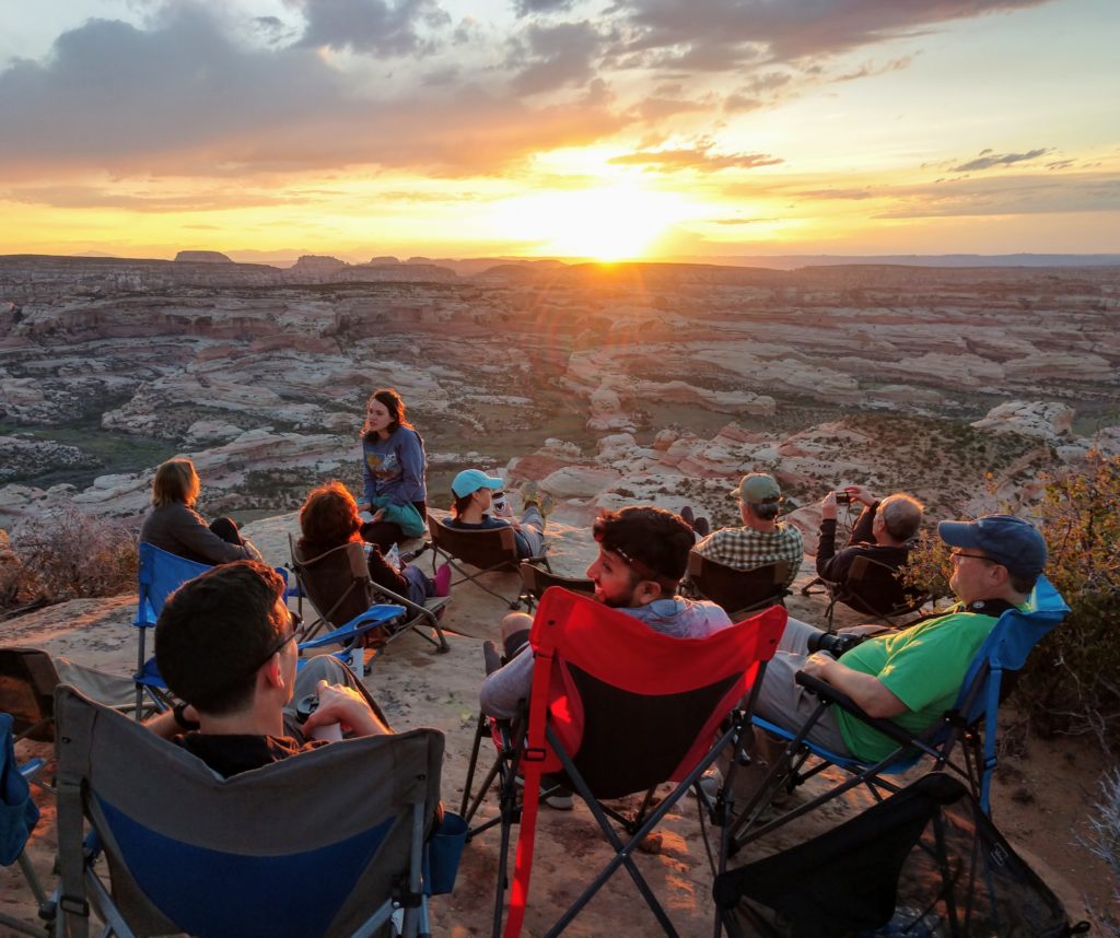 A group of cyclists relaxing and watching a sunset from on top of a bluff in Bears Ears overlooking Canyon Lands National park. As they relax in camp chairs, the Guides are busy preparing dinner.