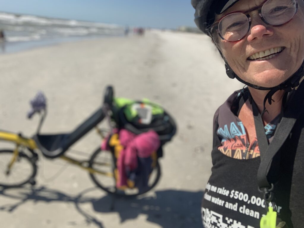 Donna taking a selfie with her bike in the back ground on the beach. 