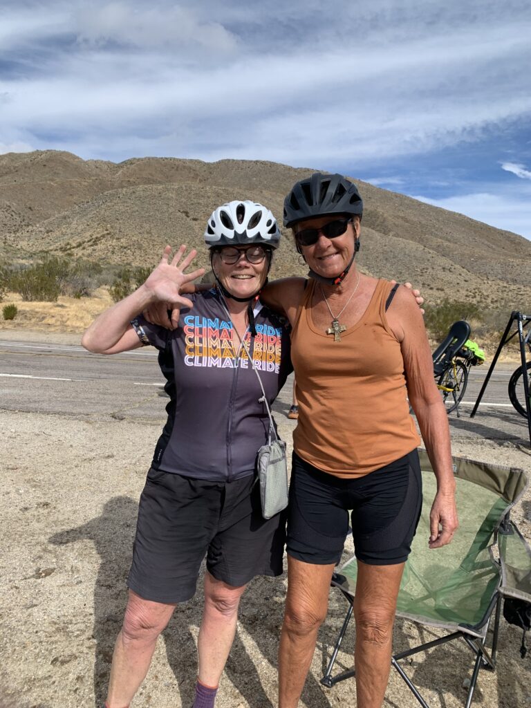 Donna and her cycling partner Becky on a previous Climate Ride. Donna is wearing a jersey on the left that reads in Climate Ride in various colors and is waving. Becky is in an orange shirt with her arm around Donna's shoulder. They are outside, pausing on a ride, their bikes behind them.