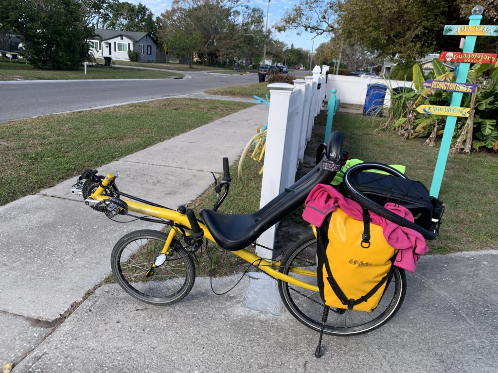 A yellow recumbent bicycle leans against a white fence, loaded up with gear for a bike packing trip.