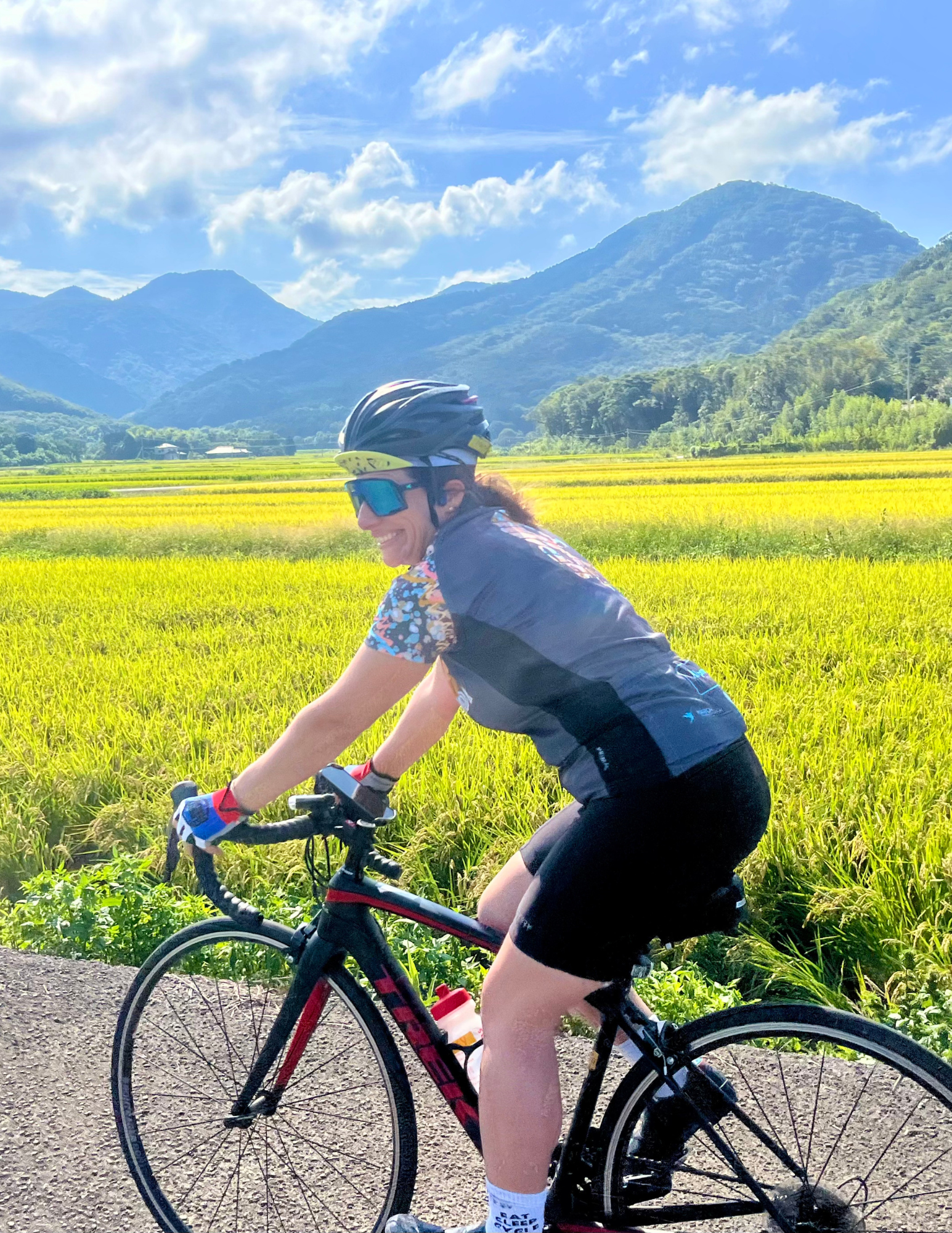 Image for the tailwinds fund which isn't a physical product. Image of a cyclist biking past a yellow field of summer flowers with mountains in the background and clouds.