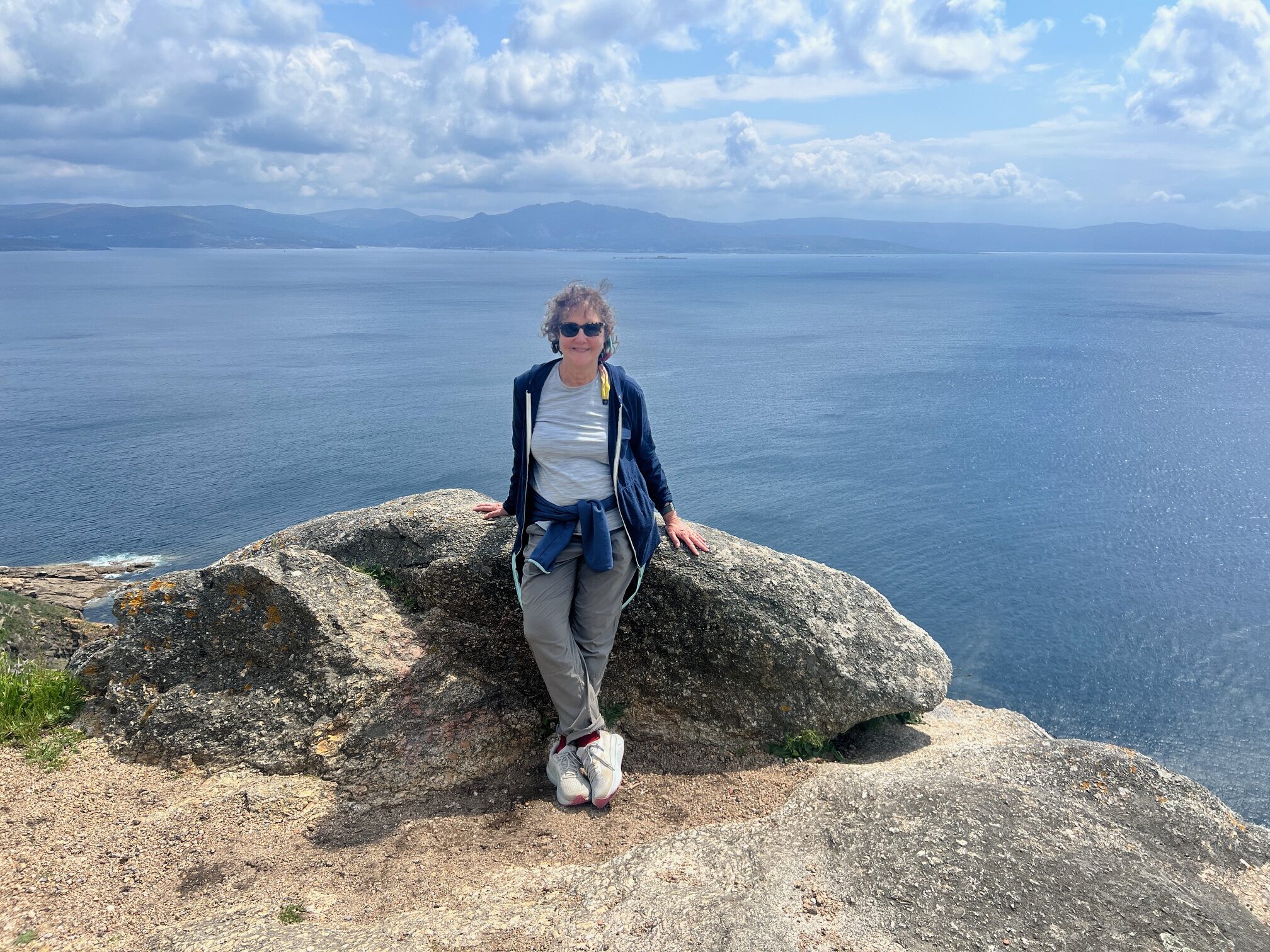 Linda Loewinger resting on the Camino against a large stone with the ocean behind her.