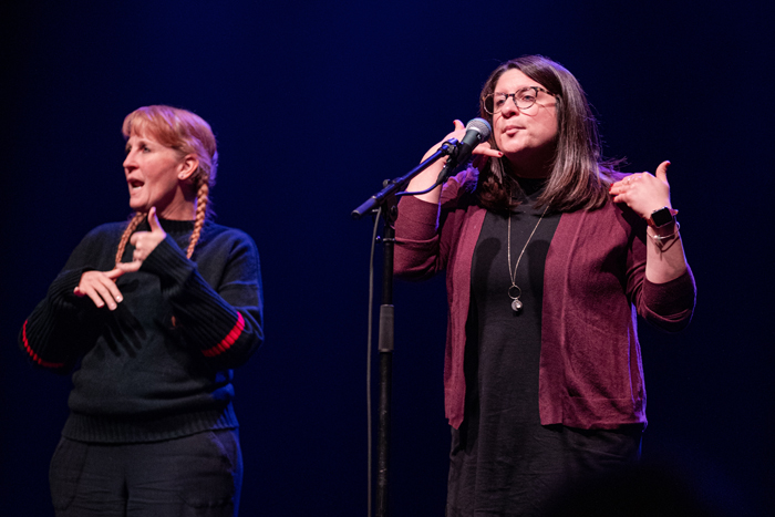 A woman shares her story on a microphone while mimicking being on a telephone. An asl interpreter stands next to her. ????kmr studios