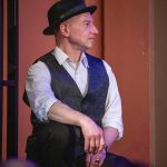 Portrait of Executive Director Marc Moss. He has a white button down shirt, a black vest and black fedora. He is seated and looking in profile away from the viewer.