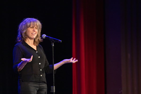 The image depicts a woman with shoulder length brown hair sharing a story in front of a microphone. There are red curtains behind her. The photo was taken by kmr studios.