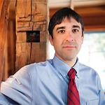 The image shows a man holding onto a wooden beam. The man is wearing a dress shirt and a tie, standing indoors against a wall.