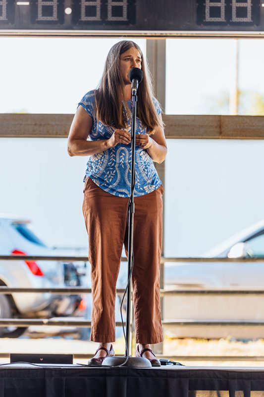 The image features a woman standing outdoors and sharing a story into a microphone. Photo credit: Donal Lakatua.