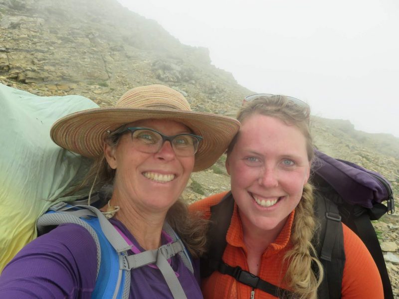 The image features a couple of women smiling for the camera outdoors. The woman on the left is wearing a sun hat. They are standing in front of a mountain backdrop.