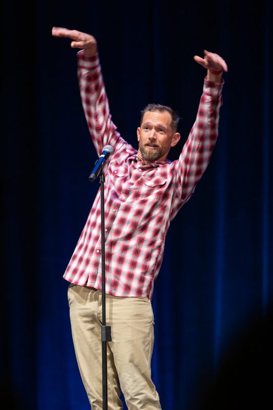 The image shows a man standing on a stage with his arms raised. The man is wearing a red plaid shirt.
