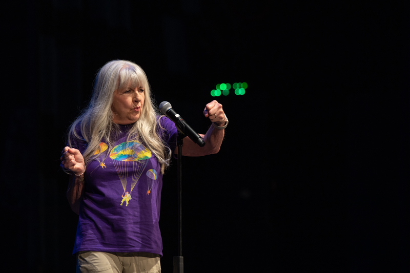 The image shows a woman standing on a stage and sharing a story. She is wearing purple clothing and appears to be performing with passion.