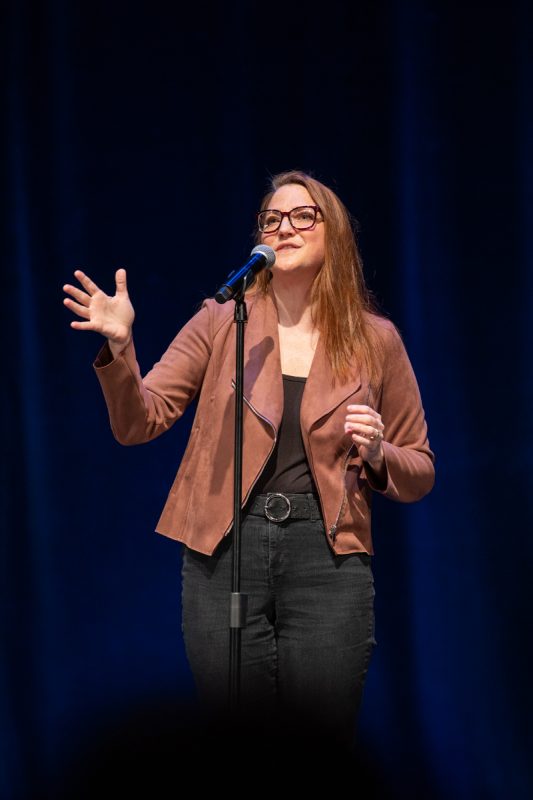 The image shows a woman with their hands up, speaking into a microphone in front of them. She is wearing a tan jacket and dark jeans.