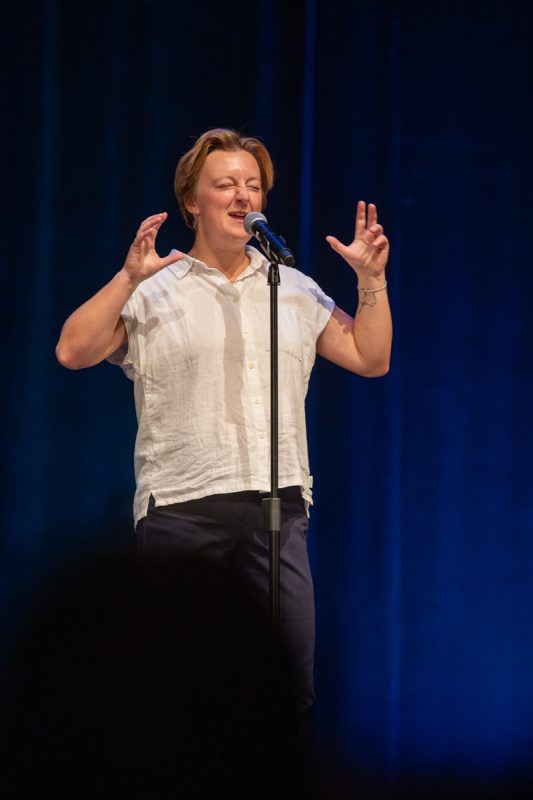 The image shows a person sharing a story into a microphone. The focus is on the individual's face as they perform, and she is very expressive. Her hands are raised in an expressive gesture. She is wearing a light-colored button-down shirt and dark pants.