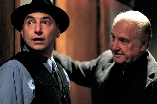 A man wearing a fedora hat is looking away from another man. The setting is indoors, and both men are dressed in fashionable clothing.