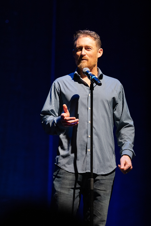 A man is standing in front of a microphone. The setting is indoors, equipped with stage equipment typically found at concerts. The photo was taken by KMR Studios.