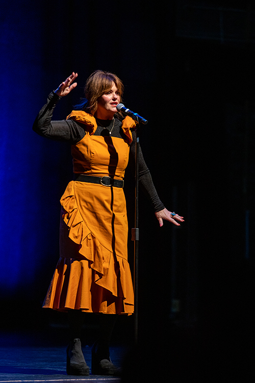 The image features a woman in an orange dress holding a microphone. She appears to be involved in a performance. The setting is indoors, emphasizing the theatrical aspect of the scene. The photo captures elements of entertainment and performing arts. Photo by kmr studios.