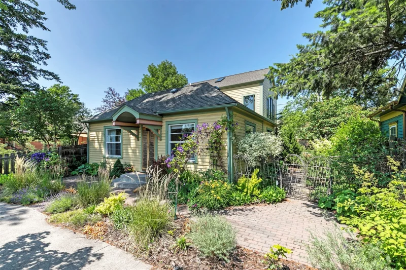 The image depicts a house surrounded by a picturesque garden. It includes elements such as trees, shrubs, and a well-maintained yard. The residence features a driveway and porch, contributing to its charming appearance within a residential area.