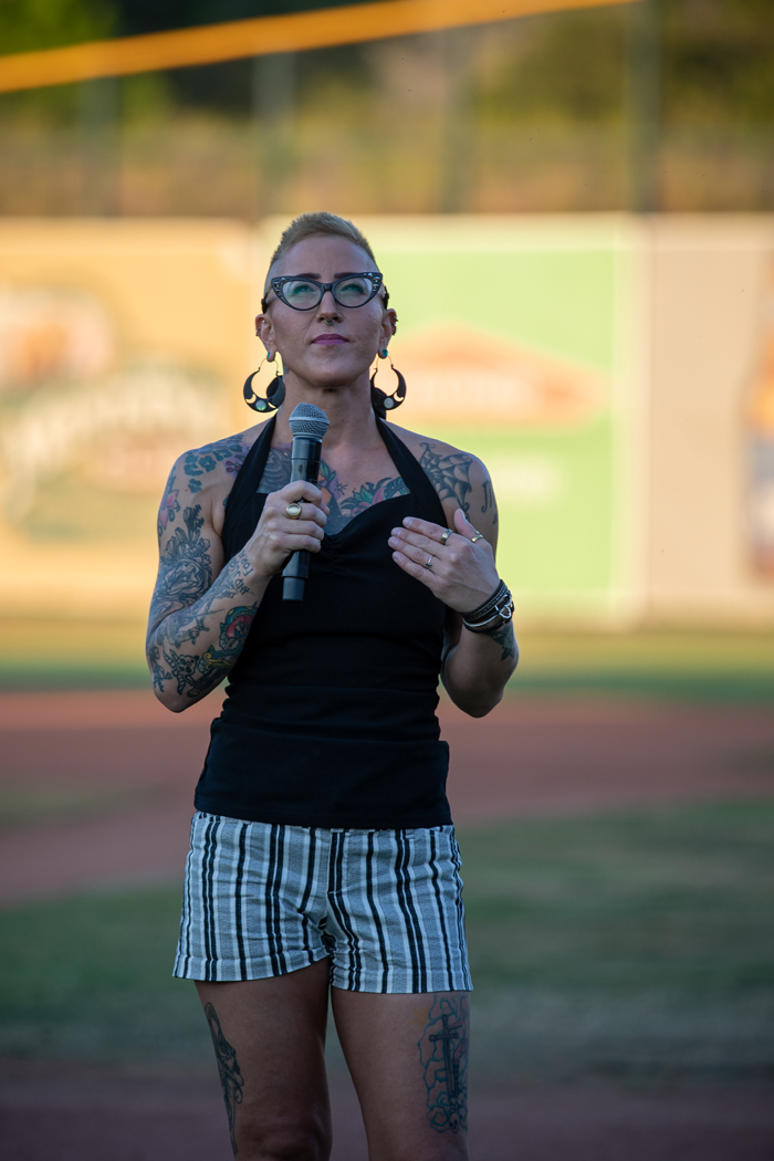 A woman is standing outdoors, holding a microphone. She is wearing a black tanktop, shorts and glasses, and the setting features grass around her. Photo credit: kmr studios