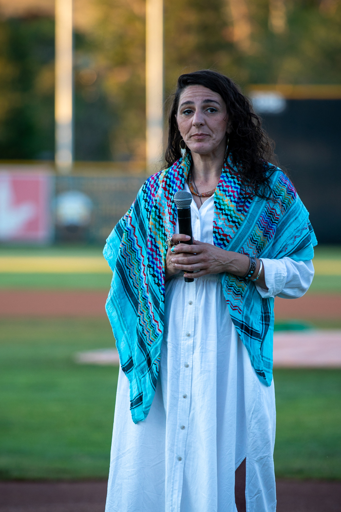A woman is standing outdoors, holding a microphone. She is dressed in fashionable clothing, and the setting features grass around her. Photo credit: kmr studios