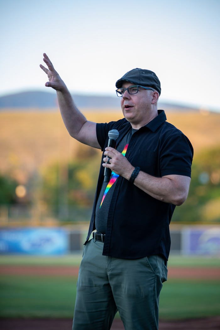 A man is holding a microphone outdoors. He appears to be dressed in clothing suitable for the environment, and there is a clear sky above. The scene suggests he may be performing, possibly related to baseball given the context of the tags. Photo credit: kmr studios