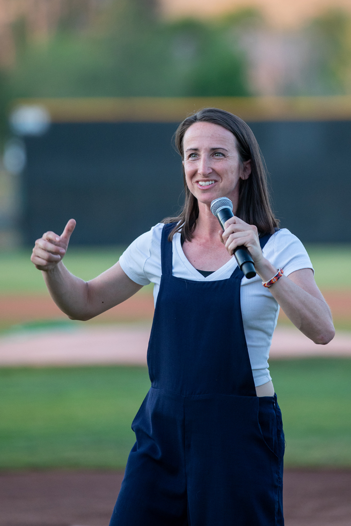 A woman is holding a microphone outdoors, likely in a grassy area. The image captures her smiling human face amidst the setting. Photo credit: kmr studios