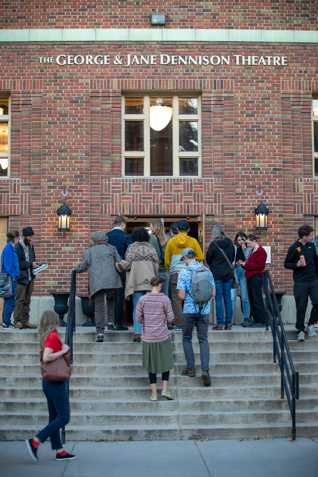 A group of people is standing on the stairs outside The George & Jane Dennison Theatre as they enter the October "Walk on the Wild Side" Tell Us Something event. They are dressed in various clothing styles, including jeans and casual footwear. The setting is outdoors in a city environment, with brick architecture visible in the background. photo credit kmr studios