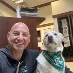 a bald smiling man poses next to a dog wearing a bandanna