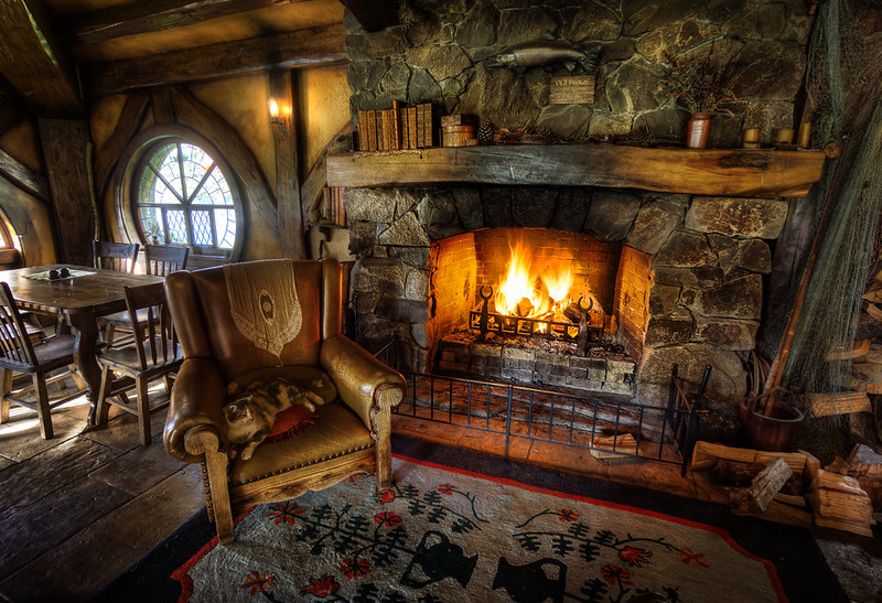 The image depicts a cozy interior of a rustic room, featuring a stone fireplace with flames flickering inside. The fireplace is adorned with wooden beams and shelves holding books. A comfortable armchair, possibly made of leather, is positioned near the fire, accompanied by an intricate rug on the wooden floor. There’s also a dining table in the background with chairs, framed by an arched window that lets in natural light. The overall atmosphere exudes warmth and comfort, reminiscent of a quaint cottage or hobbit home. Photo by Trey Ratcliff