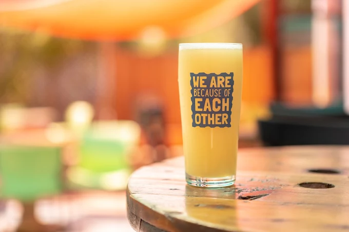 The image shows a clear glass filled with a yellow beverage, placed on a wooden table. The glass features the text "WE ARE BECAUSE OF EACH OTHER" printed on it. Soft, colorful backgrounds can be seen, suggesting an outdoor setting. Photograph by Pam Voth Photography