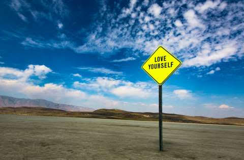 The image depicts a bright yellow caution sign with the text "LOVE YOURSELF" printed on it. The sign stands alone in a barren landscape with a clear blue sky filled with scattered clouds above and distant hills in the background.