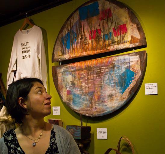 The image shows a woman looking at a large circular artwork hanging on a wall. The artwork has abstract patterns with shades of blue, red, and earthy tones, resembling natural elements like water and plants. In the background, there is also a white shirt displayed on a hanger. The wall color behind the artwork is green, contributing to the vibrant setting.