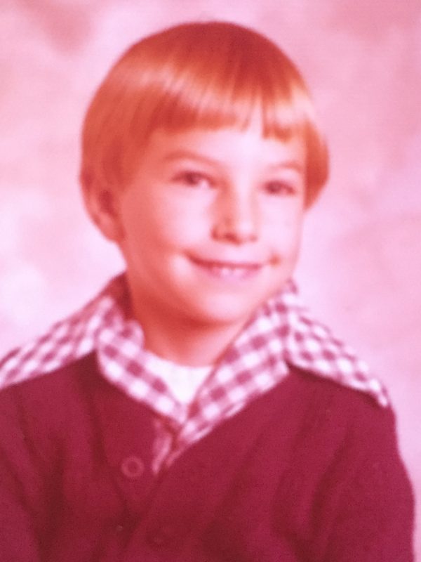 This is a vintage, 1970s-era school portrait of a young child. The image captures the quintessential aesthetic of that decade: A young boy with a classic, blondish bowl cut and a gentle, slightly mischievous smile. He is wearing a dark cardigan sweater layered over a white shirt with a wide, pointed, gingham-patterned (checkered) collar—a hallmark of 1970s children's apparel. The photo features a soft-focus studio backdrop. It has a heavy pinkish-red color cast, which is common in older color prints where the dyes have shifted and faded over time, giving it a nostalgic, warm feel.