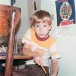 A candid shot shows a young boy in a "real-life" moment. The boy is sitting down, at a kitchen table, with his shoe propped up on his lap. He is focused on the task at hand—using a yellow plastic knife to scrape the sole of his shoe. It’s a classic "kid moment" of dealing with a messy outdoor mishap. On the wall behind him, there is a poster of a gumball machine filled with colorful characters and what looks like a hand-drawn tall figure. The wooden chair in the foreground has that heavy, dark-stained look. He is wearing a vintage-style ringer tee (white with yellow shoulders and a red collar) featuring a large number on the chest, paired with brown corduroy or cotton trousers.