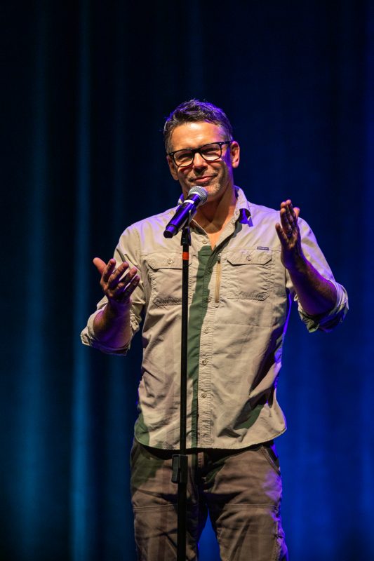 A man is standing on stage, smiling, and gesturing with his hands while speaking into a microphone. He is wearing glasses and a button-up shirt. The background has soft blue lighting, creating a relaxed atmosphere for the performance. photo by kmr studios
