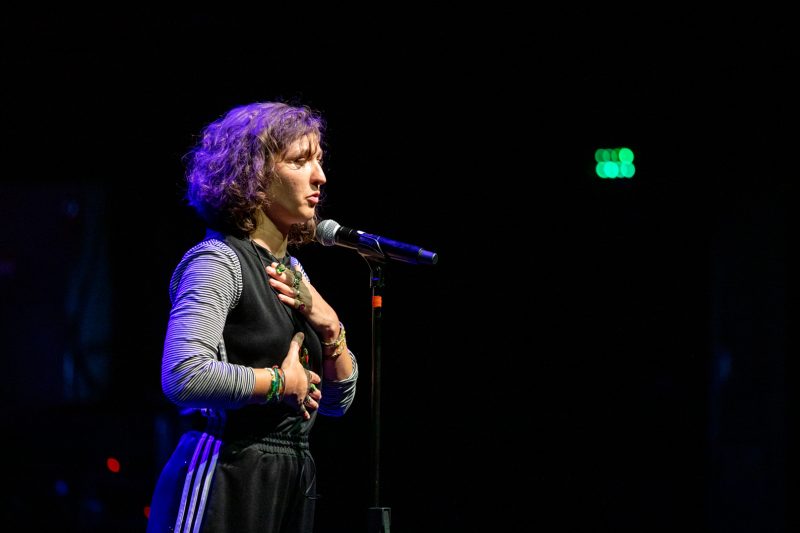 The image features a performer on stage holding a microphone. She has curly hair and is wearing a black outfit with a gray long-sleeve shirt underneath. The background appears to be dark, with soft lighting highlighting the performer. The atmosphere seems focused on her as she engages with the audience. Photo by kmr studios