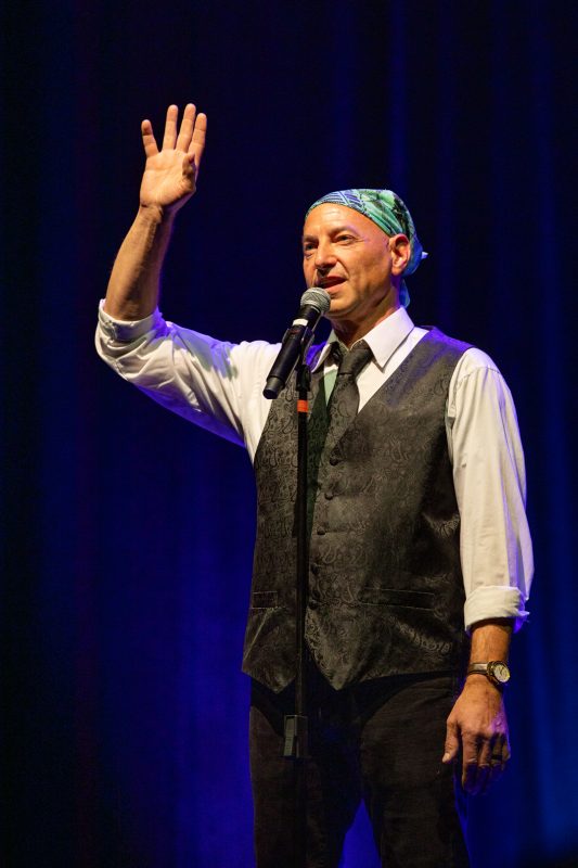 A man is standing on a stage with a microphone in hand. He is wearing a patterned bandana, a white shirt, and a dark vest. The background features soft purple lighting, creating an atmospheric setting for his performance. He appears to be engaging with the audience, raising his hand as he speaks. photo by kmr studios
