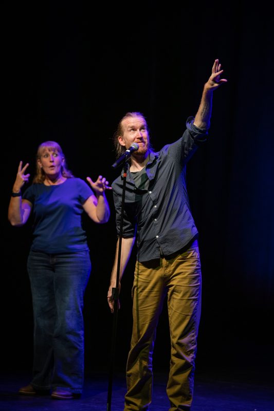 The image shows two performers on stage. One is a man with long hair gesturing expressively while speaking into a microphone, wearing a dark shirt and light-colored pants. The other performer, a woman with blonde hair, stands behind him and appears to be using sign language. The background is dimly lit, focusing on the performers. photo by kmr studios